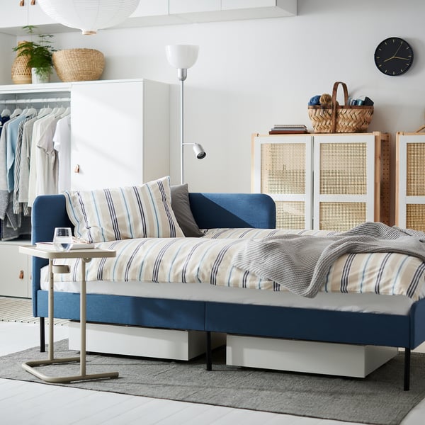 Modern, blue, convertible sofa-bed with striped cushions, white headboard, wicker baskets, and a wall clock in a minimalist room.