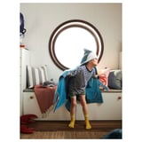 Child in blue shark towel stands before large round mirror. The towel hood, stripes, and fins. The room has a white bench, pillows, and toys.