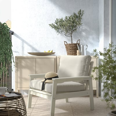 Light beige BONDHOLMEN outdoor armchair with cushions; white sideboard; basket and plant on top. Cozy balcony setting.