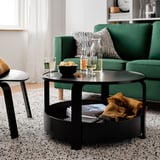Modern living room with green sofa, black coffee table holding wine glasses, snacks, and blanket. Speckled rug, white curtains.