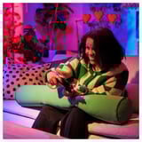 Woman using remote, sitting on colorful cushions, including a green cylindrical pillow.