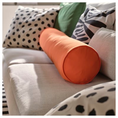 Bright orange cylindrical BRÄNNBOLL pillow on a sofa, surrounded by patterned pillows. Soft, supportive, and machine-washable.