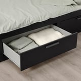 Open drawer of BRIMNES day-bed, showing folded sheets and pillows, used for storage under bed base.