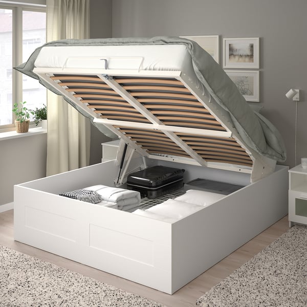 White ottoman bed with slatted base revealing storage with blankets. Near grey wall art.