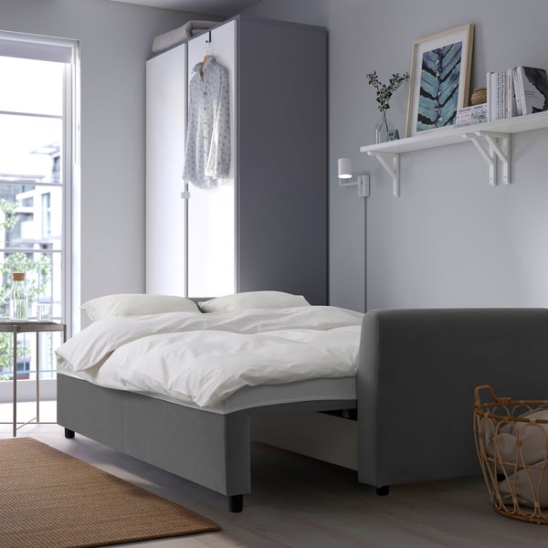 Modern minimalist bedroom with BRISSUND grey sofa bed, white bedding, hanging garment, wall shelf with decor, and natural light from window.