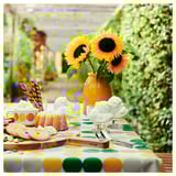 Yellow vase with sunflowers on green-yellow tablecloth. Dessert bowls, cookies, and cake.