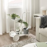 White multi-tier plant stand near light with plants, scissors, and tissues. Includes small table and grey throw.