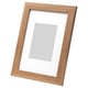 Light brown wooden picture frame, leaning against white wall, rectangular shape, neutral photo mount.
