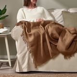 Person sitting on beige couch, wrapped in brown DYTÅG throw blanket with fringes.
