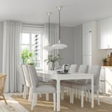 Modern white dining set with extendable table and grey chairs, large window with curtains, white pendant lights, potted plants, and kitchen cabinets.