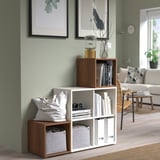Modern green living room with stacked EKET shelves holding books, bag, and decor. The shelves stand taller than the chair and near a couch.