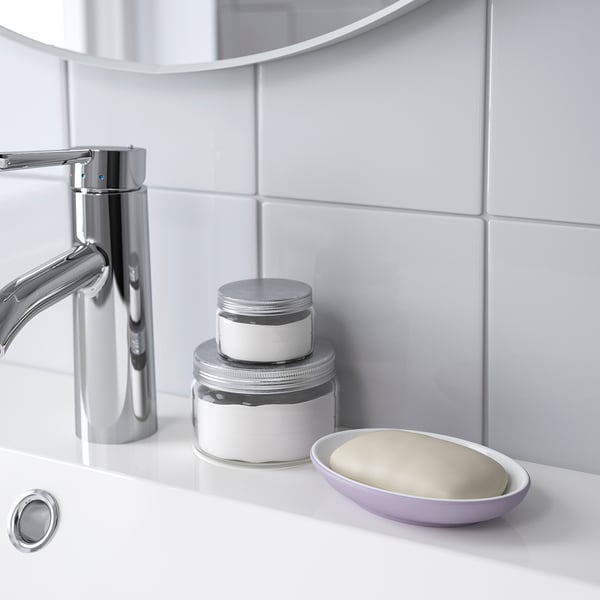 A shiny chrome faucet beside two stacked silver and white jars and a lavender soap dish with soap on a white sink.