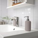 Modern bathroom with white countertop, shiny chrome faucet, pinkish-grey soap dispenser, small plant, and toothbrush holder; clean, minimalist design.