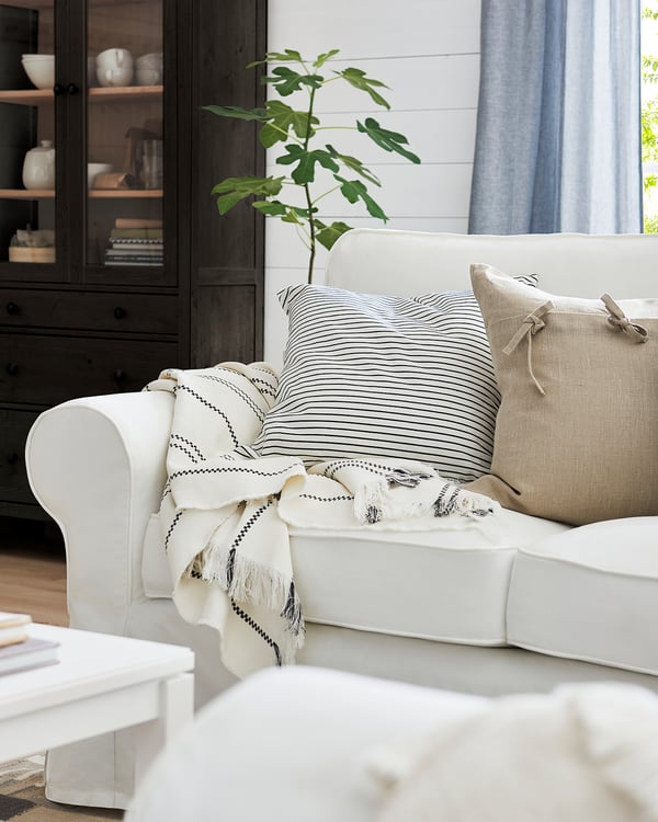 White EKTORP sofa with striped pillows and a throw blanket in a cosy room. Dark cabinet and plant near window with blue curtains.