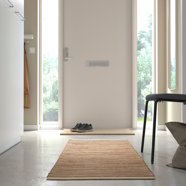 A minimalist hallway with a beige doormat, black sneakers, and a grey chair. A handwoven rug decorates the polished floor.