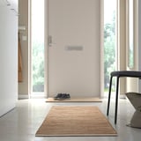 A minimalist hallway with a beige doormat, black sneakers, and a grey chair. A handwoven rug decorates the polished floor.