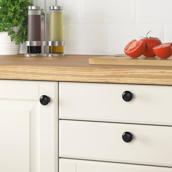 Wooden kitchen counter with white drawers featuring ENERYDA black knobs. Nearby, there are colorful bottles, fresh tomatoes, and a plant.