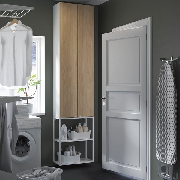 ENHET storage with doors & shelves holding laundry items & plant, beside ironing board.