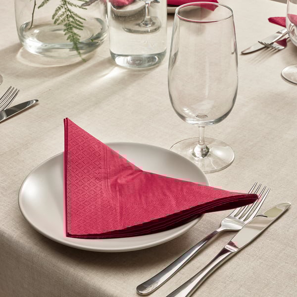 Dining table set with white plate, red napkin, glass, fork, knife, beige cloth, and vase with greenery.