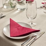 Dining table set with white plate, red napkin, glass, fork, knife, beige cloth, and vase with greenery.