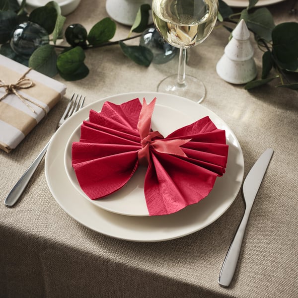 A table set with a white plate, red folded napkin, fork, knife, wine glass, green leaves, and small gift.