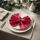 A table set with a white plate, red folded napkin, fork, knife, wine glass, green leaves, and small gift.