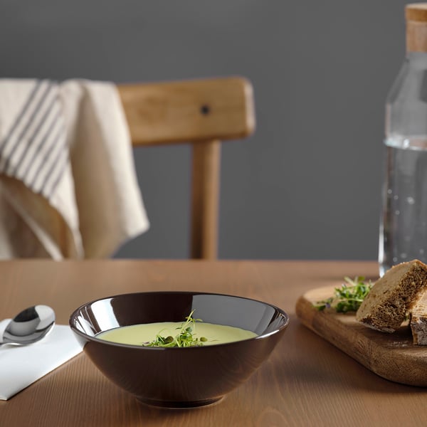 Black bowl with creamy soup, spoon, napkin, bread, chair, and water bottle on a wooden table.