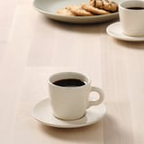 A small white cup of coffee on a saucer, paired with another cup and plate of cookies, on a wooden table.