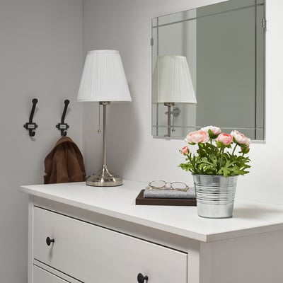 Small white potted artificial plant with pink flowers on a dresser near lamps.