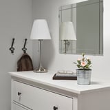 Small, gray-potted artificial pink flowers on a white dresser, flanked by lamps and a mirror.