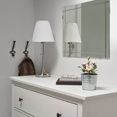 Small, gray-potted artificial pink flowers on a white dresser, flanked by lamps and a mirror.