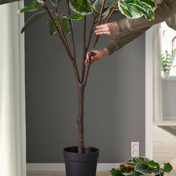 Person touches potted artificial plant in modern room. Its suitable for indoor and outdoor use, providing year-round greenery without maintenance.