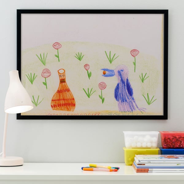 Childs drawing in black frame on white desk. Left lamp illuminates artwork and school supplies.