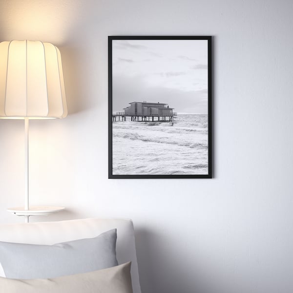 Black FISKBO frame on wall, displaying pier scene, above couch, next to lamp.