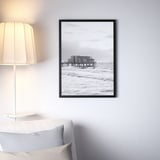 Black FISKBO frame on wall, displaying pier scene, above couch, next to lamp.