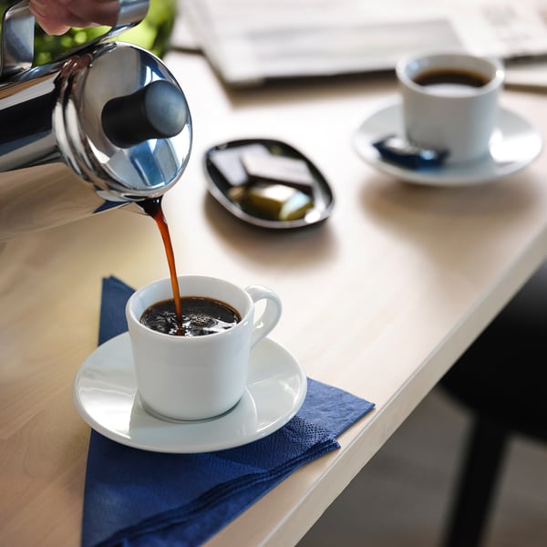 Person pouring coffee from metal pot into white cup on saucer and napkin. Another cup of coffee nearby.