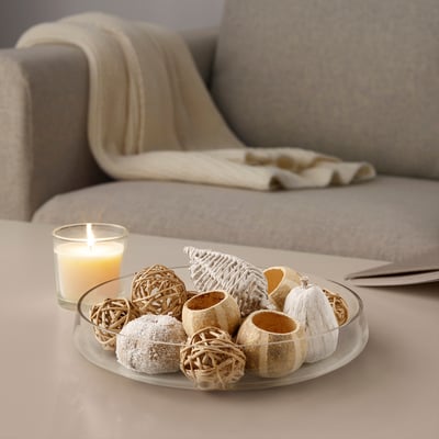 Glass bowl holds decorative potpourri items, including a lit candle, near a beige couch.