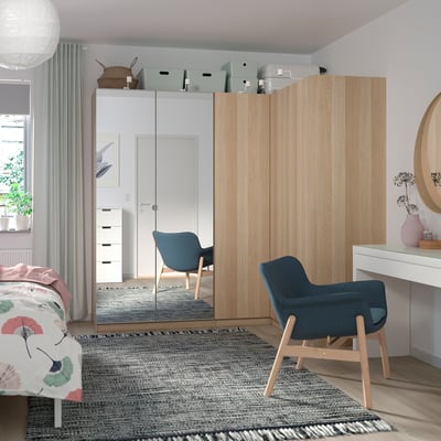 Bedroom with light wooden wardrobe, large mirror, blue chair, white desk, rug, and bed with colorful blanket.