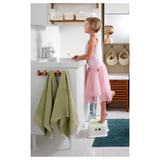 A girl in a pink dress uses a white FÖRSIKTIG stool to reach a bathroom sink. The stool has an anti-slip top and steady base for safety.