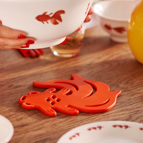 Red FÖSSTA trivet with goldfish design on wooden table.
