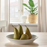 A bowl of green pears sits on a wooden table near a window with a potted plant.