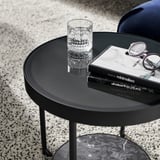 Black circular side table with glass top, metal legs, and marble-effect shelf. A glass cup, book, and glasses placed on it.