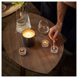 Person lights candles on wooden table: large scented and small tea lights, empty glasses, vase with plant. Cozy lighting.