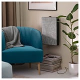 A cozy blue FULLÖSA armchair sits in a living space, featuring a gray throw blanket and accompanied by a stack of magazines and a potted plant.