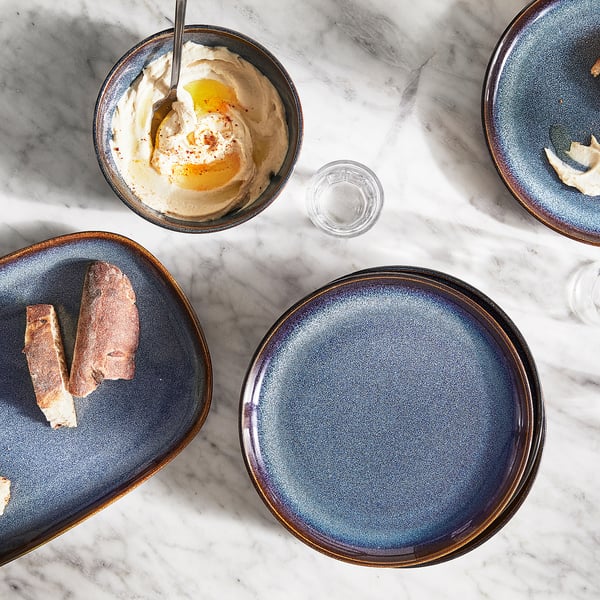 Blue glazed stoneware bowl, golden rim, oval shape. Table setting with white food, marble surface.