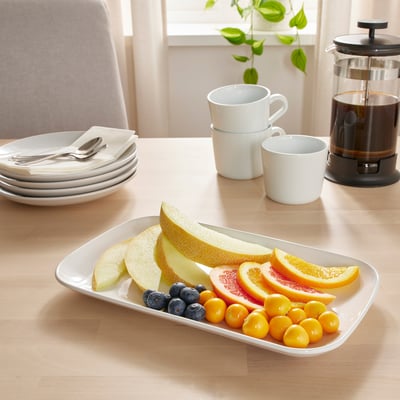 Platter filled with honeydew, cantaloupe, orange slices, blueberries, and tomatoes near coffee mugs, plates, and a french press.