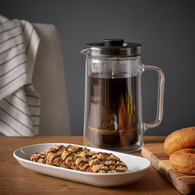 Clear coffee press on wooden table, beside it a plate of chocolate chip cookies and sugared donuts.