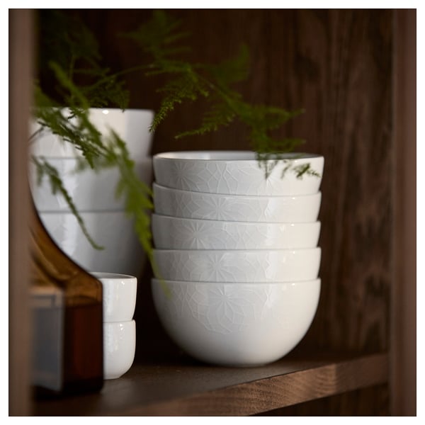 Stack of six white GOKVÄLLÅ bowls with embossed geometric patterns on wooden shelf, next to green plant.