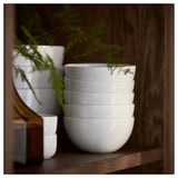 Stack of six white GOKVÄLLÅ bowls with embossed geometric patterns on wooden shelf, next to green plant.
