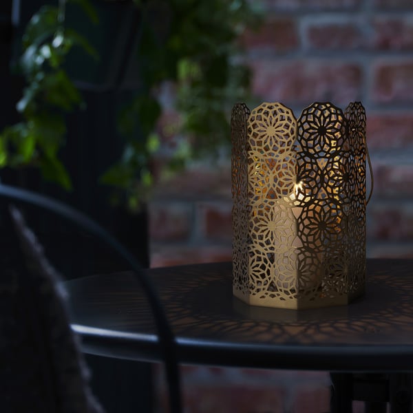 A decorative lantern sits on an outdoor table, emitting a warm glow through its intricate floral pattern. The long handle allows for easy carrying.